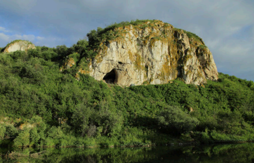 a picture of a cave in the edge of a cliff overlooking a forest and a river