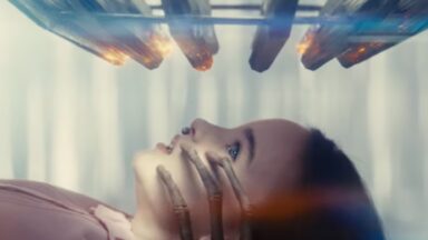 little girl lying on her back in a clinical setting looking up at an array of rods and lights comes down toward her. Spindly alien fingers are touching her face from below.