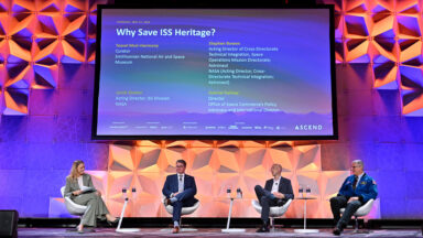 Four people sit on a colorfully-lit stage under a large screen displaying their panel title, names and titles.