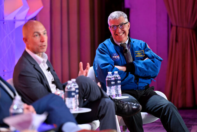 Two men, including an astronaut wearing a blue flight jacket, sit on a colorfully-lit stage during a panel discussion.