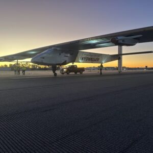 A predawn image of the Skydweller Aero drone with jumbo jet sized wings sitting on a runway or taxiway. Three people stand near the aircraft's right wing while another person is walking near the left wing. A pickup truck is parked alongside the aircraft.