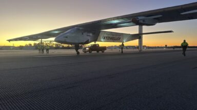 A predawn image of the Skydweller Aero drone with jumbo jet sized wings sitting on a runway or taxiway. Three people stand near the aircraft's right wing while another person is walking near the left wing. A pickup truck is parked alongside the aircraft.
