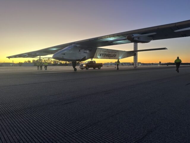 A predawn image of the Skydweller Aero drone with jumbo jet sized wings sitting on a runway or taxiway. Three people stand near the aircraft's right wing while another person is walking near the left wing. A pickup truck is parked alongside the aircraft.
