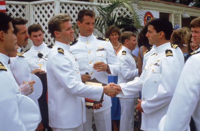two naval officers in white dress uniforms shaking hands while several others look on