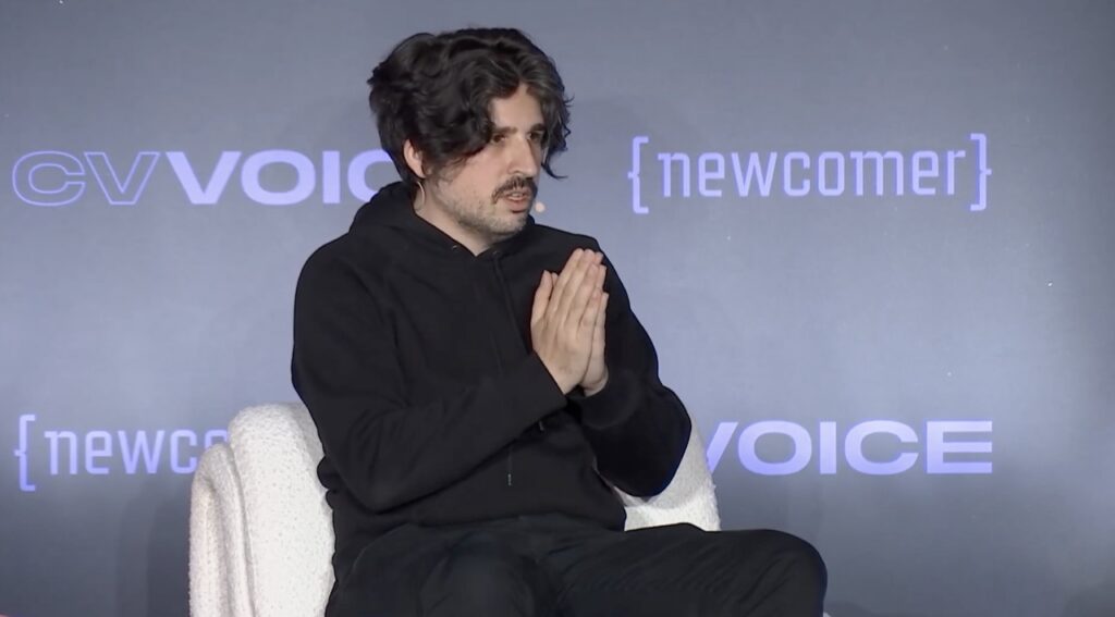 A man in a black hoodie gestures as he speaks on stage
