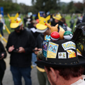 A player wearing a hat decorated with Pokemon characters and trading cards plays Pokemon GO on a smartphone during the in-person Pokemon GO Tour: Kalos Los Angeles 2026 event at the Rose Bowl Stadium in Pasadena, California, on February 20, 2026.