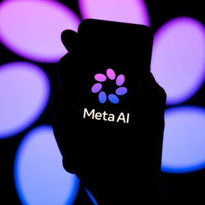 The words "Meta AI" in white are seen with a purple petal pattern logo for Meta AI on a dark smartphone screen. A dark hand holds the phone. The background shows a bigger version of the Meta AI logo.