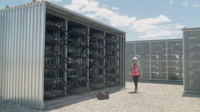 A woman wearing a white safety helmet and a red safety vest is walking by a large steel structure containing used EV battery packs connected together. Such repurposed EV batteries provide 32 megawatt-hours of energy storage to support the local grid in Lancaster, California.