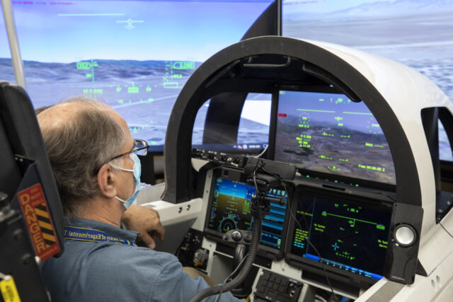 A close up of the cockpit view of the eXternal Vision System before it was placed in the X-59. Instead of a front facing window, the pilot uses monitors for forward facing visibility. A man wearing a surgical mask sits inside the cockpit mockup.