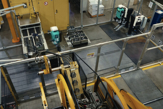 Two Digit humanoid robots developed by Agility Robotis are seen working in an isolated "work cell" within a factory.