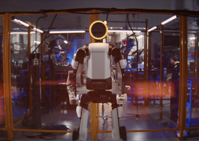 An Atlas humanoid robot made by Boston Dynamics stands with arms at its sides. The background shows a caged area separating the robot from human researchers.