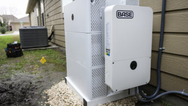 A white-colored residential battery with the name "BASE" on the side sits on the side of a brown-colored house in Houston, Texas.
