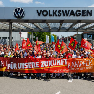 Employees of German car maker Volkswagen (VW) wave flags and display a banner with the lettering 'To fight united for our future - all workforces across the VW Group' as they take part in a rally called by German trade union IG Metall to protest against restructuring and mass job cut plans at the VW plant in Zwickau, eastern Germany on July 9, 2026. IG Metall is organising protests by VW workers outside plants across the country on July 9, when the carmaker's bosses will present the restructuring plans to the supervisory board. Europe's largest carmaker has come under intense pressure from US tariffs, slimmer profit margins from electric cars and above all intense competition in China, the world's largest auto market. VW, whose 10 brands range from mass-market Seats to premium Porsches, is already in the process of axing 50,000 jobs in Germany by 2030, including 35,000 at its namesake marque. (Photo by Jens SCHLÜTER / AFP via Getty Images)