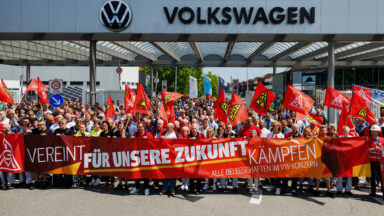 Employees of German car maker Volkswagen (VW) wave flags and display a banner with the lettering 'To fight united for our future - all workforces across the VW Group' as they take part in a rally called by German trade union IG Metall to protest against restructuring and mass job cut plans at the VW plant in Zwickau, eastern Germany on July 9, 2026. IG Metall is organising protests by VW workers outside plants across the country on July 9, when the carmaker's bosses will present the restructuring plans to the supervisory board. Europe's largest carmaker has come under intense pressure from US tariffs, slimmer profit margins from electric cars and above all intense competition in China, the world's largest auto market. VW, whose 10 brands range from mass-market Seats to premium Porsches, is already in the process of axing 50,000 jobs in Germany by 2030, including 35,000 at its namesake marque. (Photo by Jens SCHLÜTER / AFP via Getty Images)