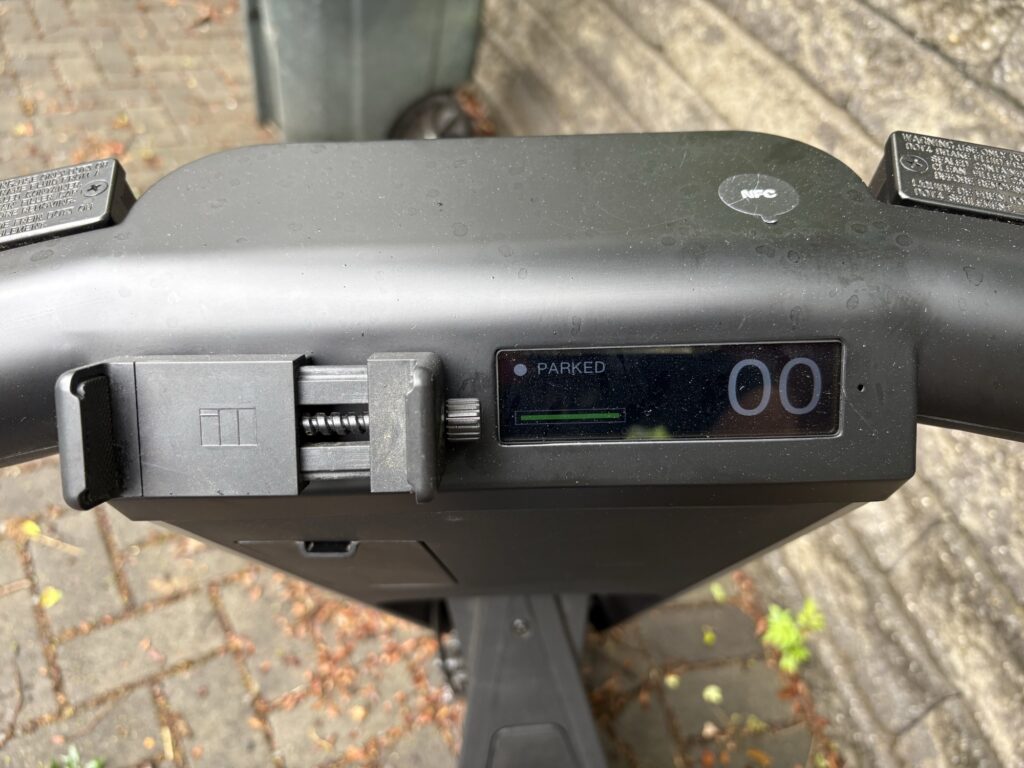 Image of the top of a scooter's handle bars, showing a small display and a large clamp for holding a phone.