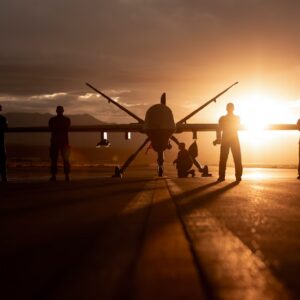 432nd Wing/432nd Air Expeditionary Wing Airmen pose with an MQ-9 Reaper for a photo at Creech Air Force Base in Nevada on Nov. 19, 2019. The drone is seen silhouetted against the rising or setting sun with six people standing in front alongside its wings, and one person crouched near its landing gear.