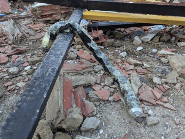 A long black snake robot covered in plastic material lies on top of a bar amid building debris from an earthquake.