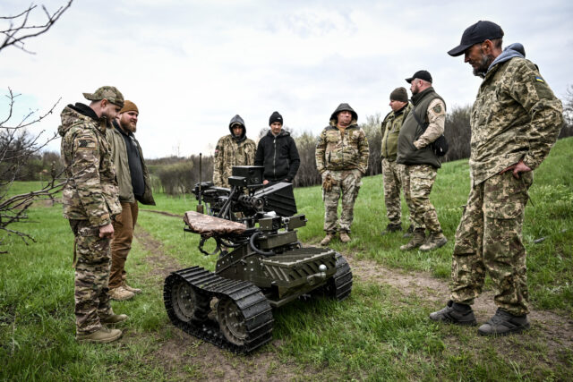 Ukrainian soldiers stand around a ground robot that is armed with a machine gun.