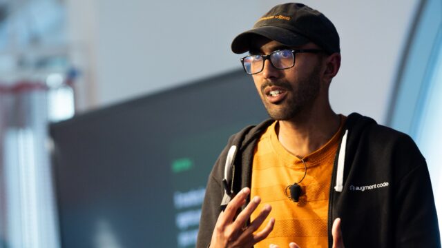 A man with a baseball cap and glasses gestures and speaks on a stage