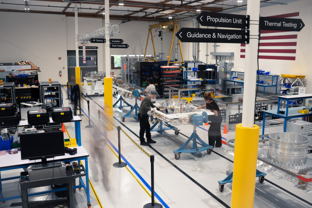 A view of the Xona Space Systems satellite factory in Burlingame, California. The foreground shows a signpost with signs pointing toward "propulsion unit," "guidance & navigation" and "thermal testing."