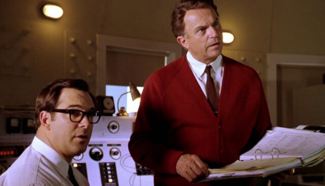 two scientists one sitting, one standing, the latter wearing a red sweater over a dress shirt and tie, with communications equipment behind them
