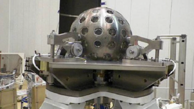 Image of a spherical silver object with regularly spaced features on its surface. It's he'd in a large room by a complicated support structure.