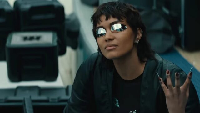 A girl with short dark hair, wearing futuristic glasses and raising a hand with long metallic nails
