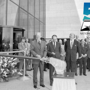 A black and white photo of a crowd waiting and watching for a robotic arm to snip a ribbon outside of a building
