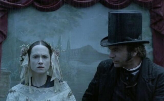 man and woman in period wedding garb posing for a photograph, she looking straight ahead, he looking at her