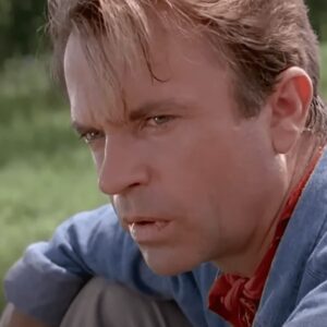 ruggen paleontologist in blue shirt and red kerchief looking intently off to the side while sitting in a jungle clearing