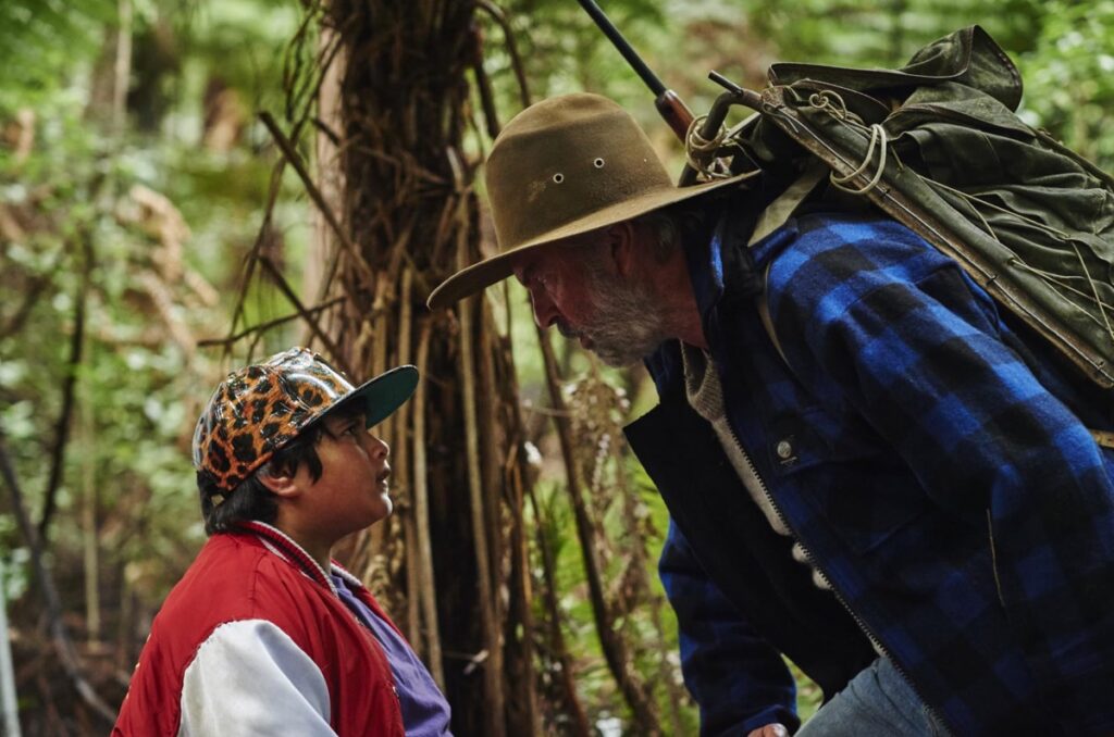 plump young boy in baseball cap and red and white jacket facing off against older man in hunting gear in the woods