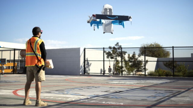 An Amazon Prime Air drone hovers above a paved lot in front of a person wearing an orange safety vest.