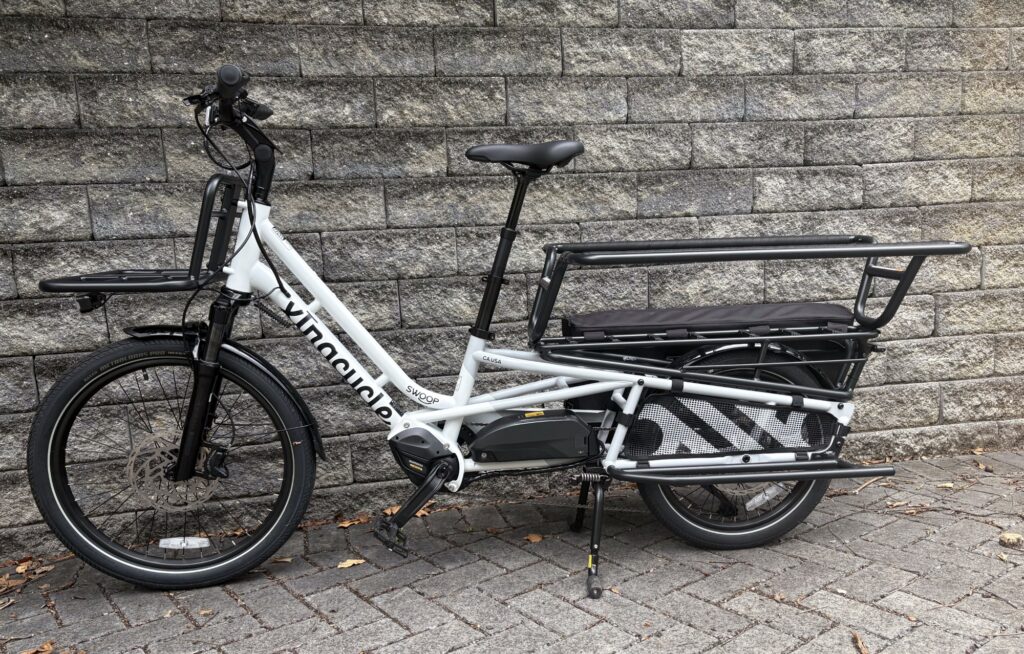 Image of a white bicycle with black handlebars and cargo rack. The rear wheel of the bicycle is on an extended frame.