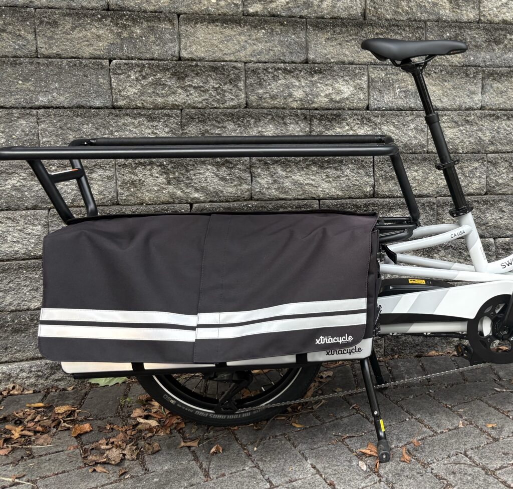 Image of the rear of a long-tail cargo bike, showing metal hand rails extending up. There is a large bag attached to the frame under the seating area.