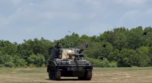Quadcopter drones equipped with Auterion's Nemyx swarm autonomy system crash into a self-propelled gun sitting in a field with trees in the background during a test demonstration.