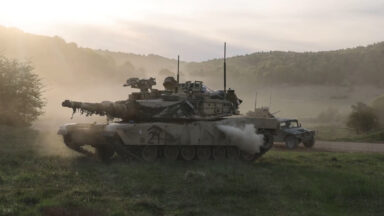 A US Army tank can be seen in profile with its main gun facing to the left. The background shows another US Army vehicle driving by with dust in the air. The scene took place during Exercise Combined Resolve held in Germany from April 9 to May 10, 2026.