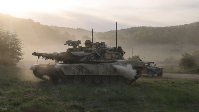 A US Army tank can be seen in profile with its main gun facing to the left. The background shows another US Army vehicle driving by with dust in the air. The scene took place during Exercise Combined Resolve held in Germany from April 9 to May 10, 2026.