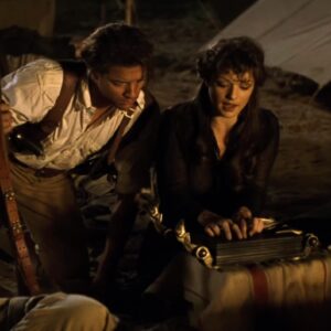 man in 1920s adventurer garb crouched near a woman beginning to read an ancient book at a desert camp site