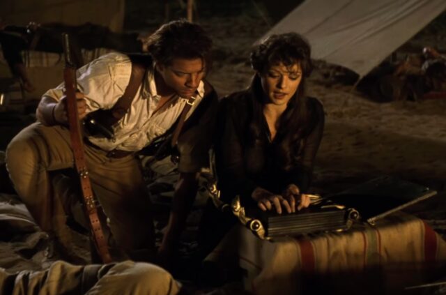 man in 1920s adventurer garb crouched near a woman beginning to read an ancient book at a desert camp site