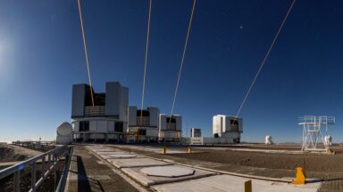 Image of four cylindrical buildings, each with an open slot for the telescope, and each emitting an orange colored laser