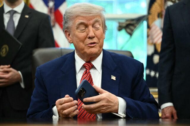 President Trump sitting at his desk in the White House while holding a smartphone in his left hand.