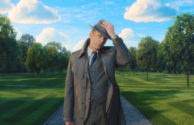 still image showing a man in great raincoat and hat standing against an idyllic background of green flora and blue skies