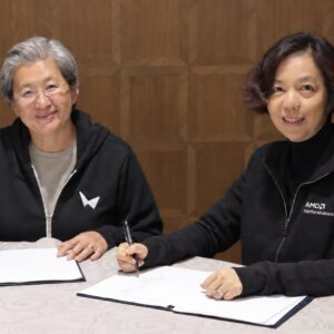 Two women in black hoodies—one from AMD wearing a World Labs hoodie, and one from World Labs wearing an AMD hoodie, smile at the camera while sitting a desk with papers to sign on it.