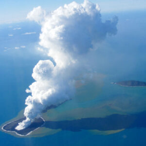 A pillar of white cloud arises from a small island in the ocean.