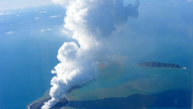A pillar of white cloud arises from a small island in the ocean.