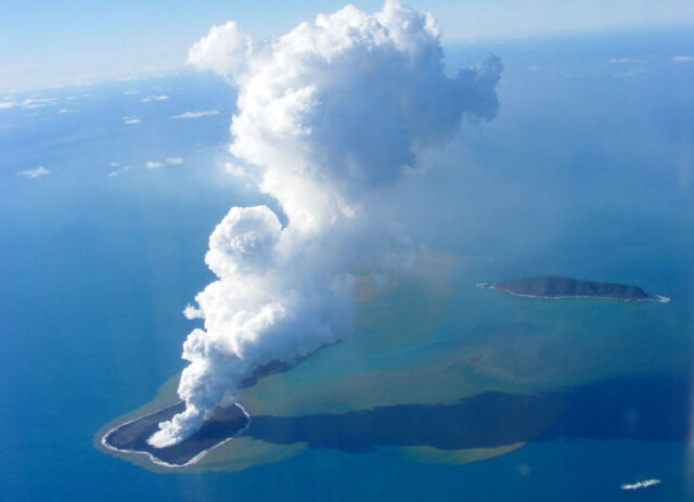 A pillar of white cloud arises from a small island in the ocean.