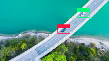 two cars drive on a bridge, seen from above. One is bounded by a green box, one by a red box