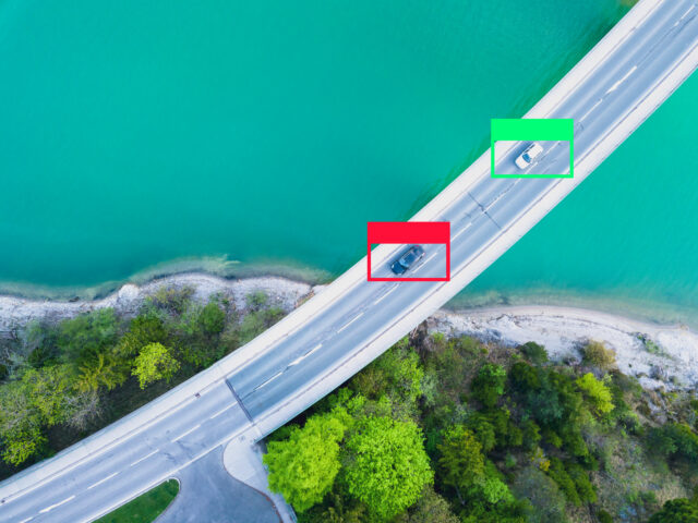 two cars drive on a bridge, seen from above. One is bounded by a green box, one by a red box