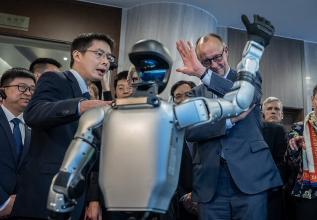 Unitree Robotics founder Wang Xingxing gives German Chancellor Friedrich Merz a tour on February 26, 2026 in Hangzhou, China. Wang is seen standing on the left with his hand on the shoulder of a waving humanoid robot, while Merz is seen standing on the right with his left arm raised to wave back to the robot.