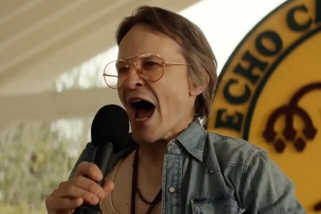 middle aged man with sandy hair and glasses speaking loudly into a handheld microphone